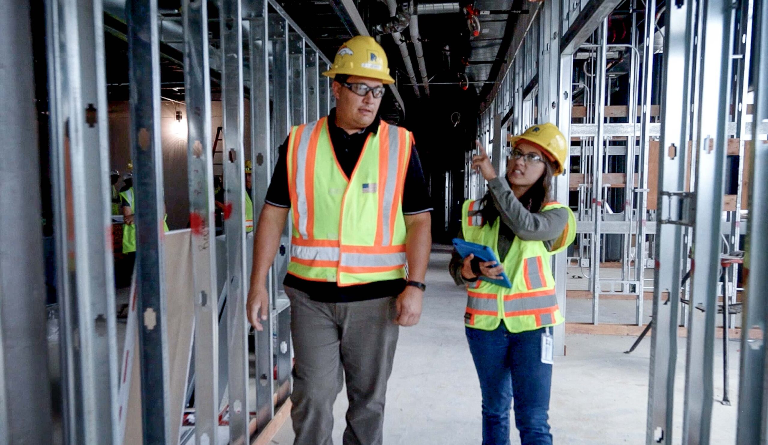 Man and woman walking through a construction site wearing a hard hat and bright yellow vests
