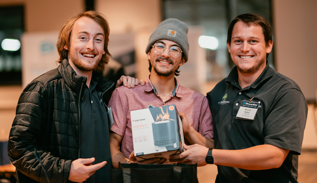 Three young men smiling holding a stainless steal bonfire gift