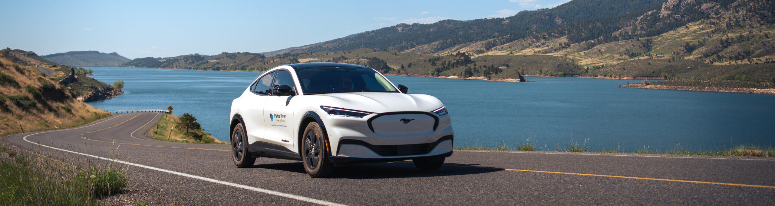 White electric vehicle driving on a road with a lake and mountains in the background.