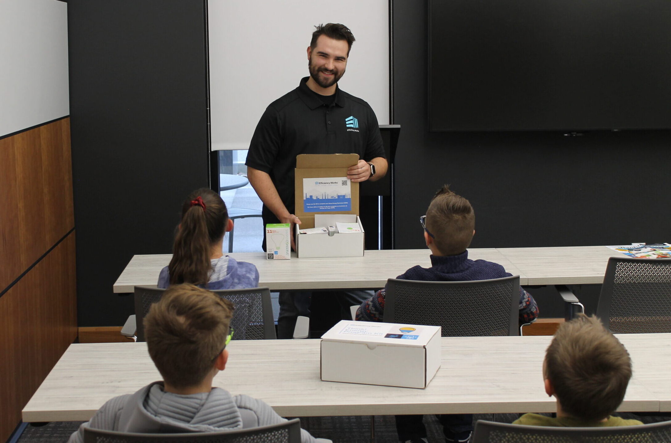 Photo of a young man opening an Efficiency Works kit showing it to four kids sitting at their tables.