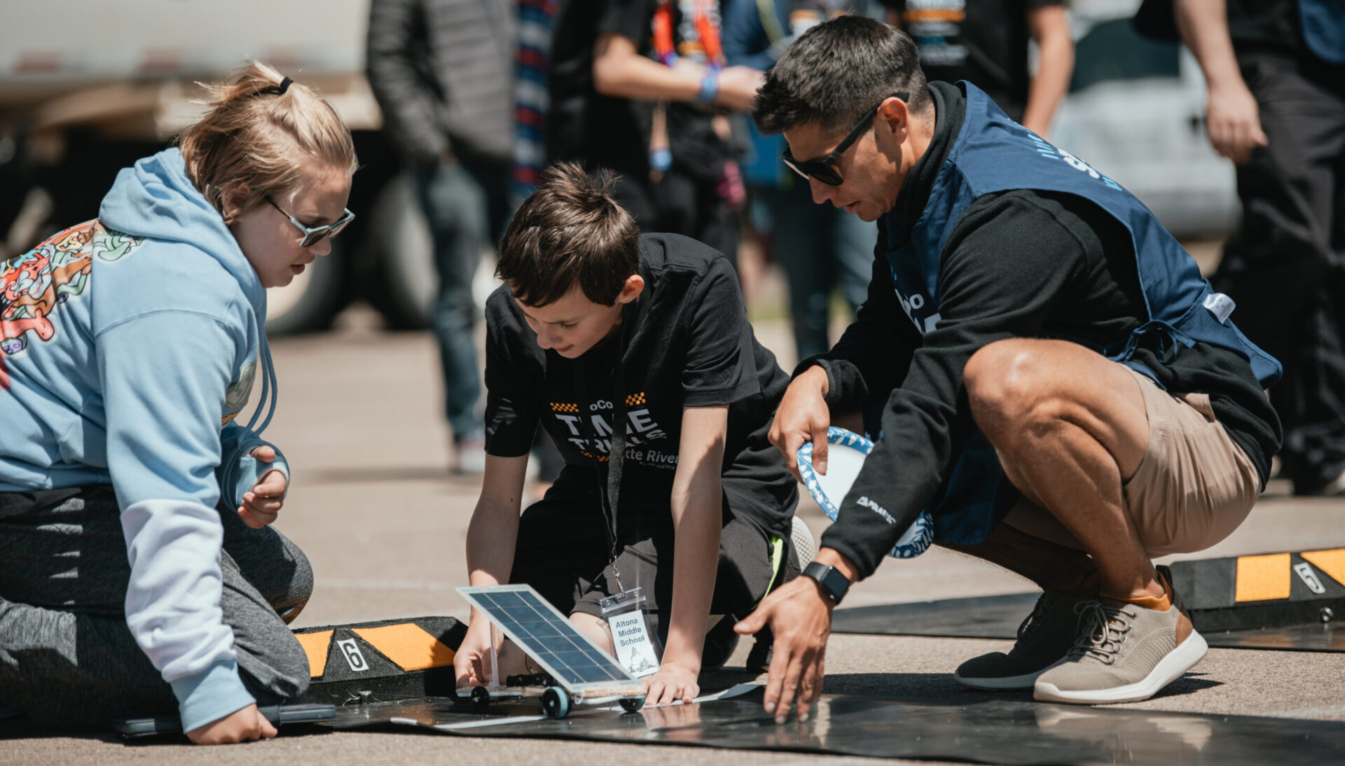 Young man helping two kids set up a solar powered car at an event.