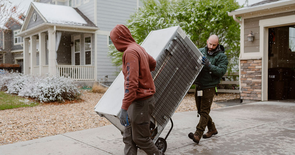 Two men hauling away a white old refrigerator from a home while it snows outside