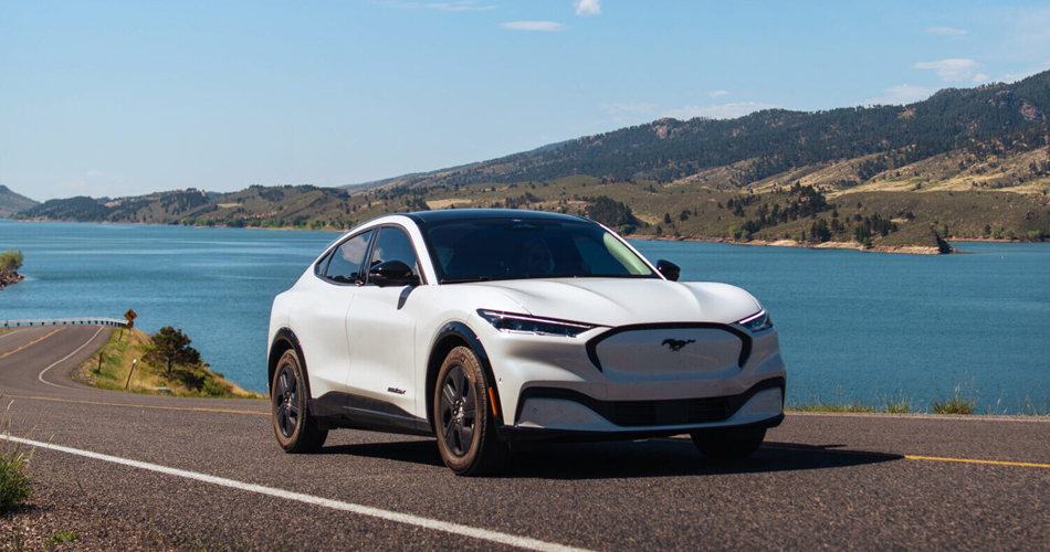 White electric vehicle driving on a road with a lake and mountains in the background.