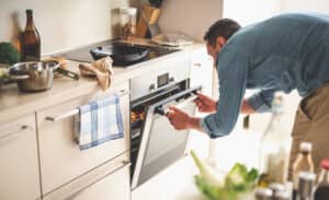 Portrait of gentleman in denim shirt closing door of oven while looking at frying pan with beef steak
