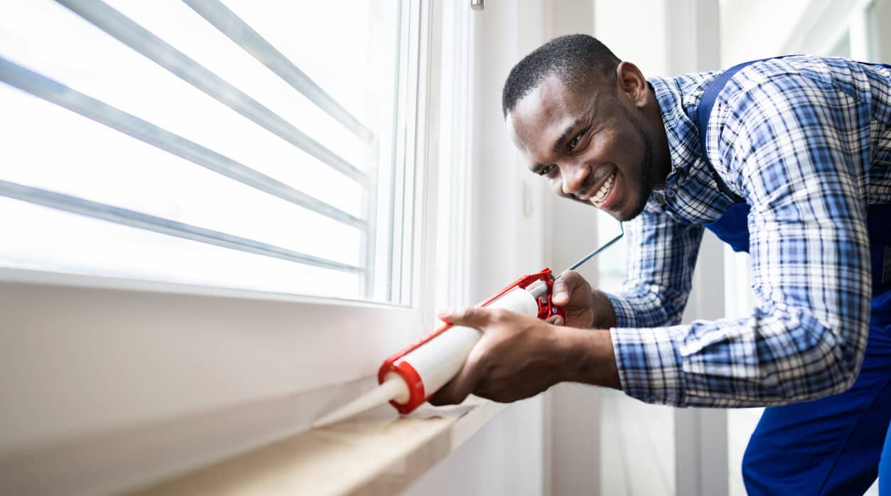 Young African American man applying caulk sealant to window interior