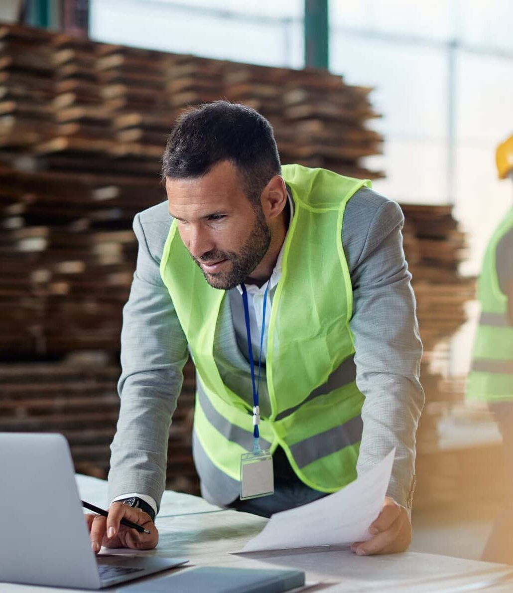 Warehouse manager working on laptop while going through paperwork at lumber distribution department.
