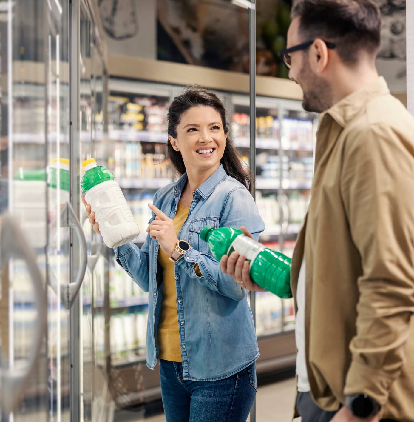 A smiling couple is choosing between milk and yogurt while standing at dairy section in supermarket.