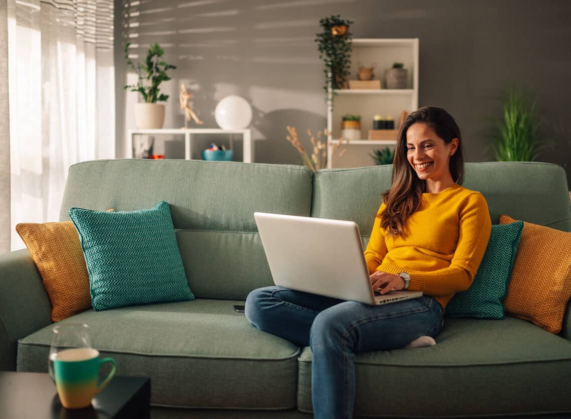 Smiling woman using laptop while sitting on a mint couch at home.
