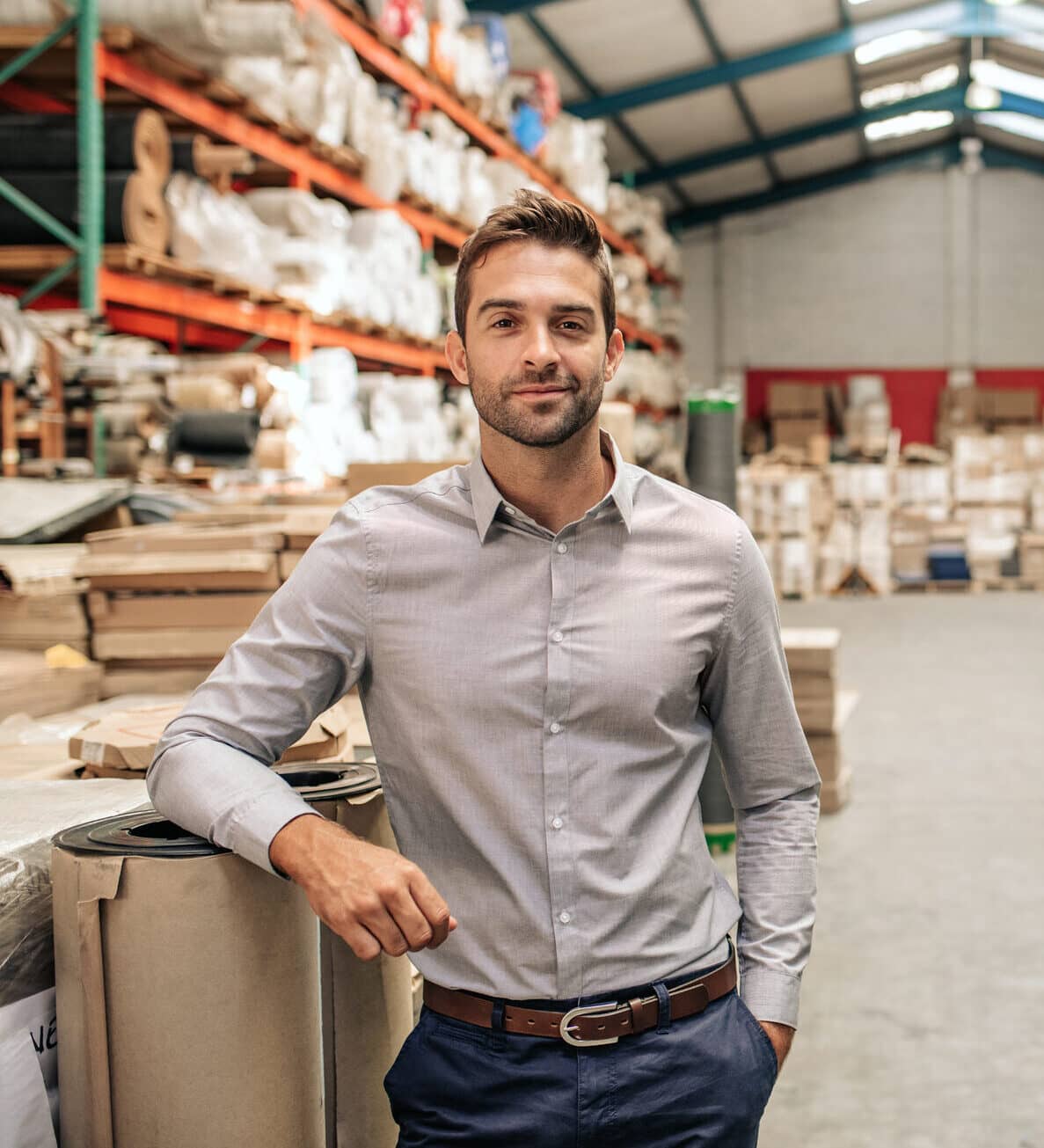Portrait of a smiling warehouse manager leaning against some stock with piles of carpets on shelves and a forklift in the background