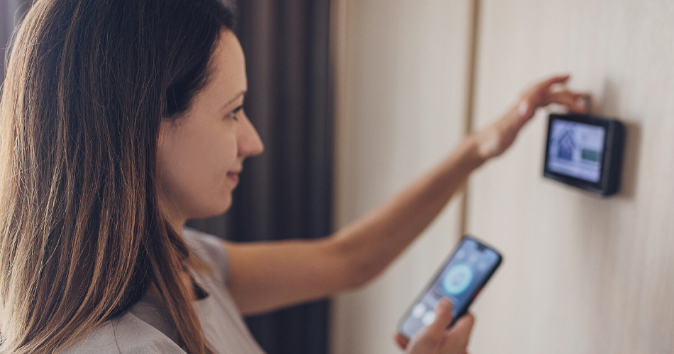 Close up of a woman holding a smart phone in one hand and adjusting a smart thermostat with the other.