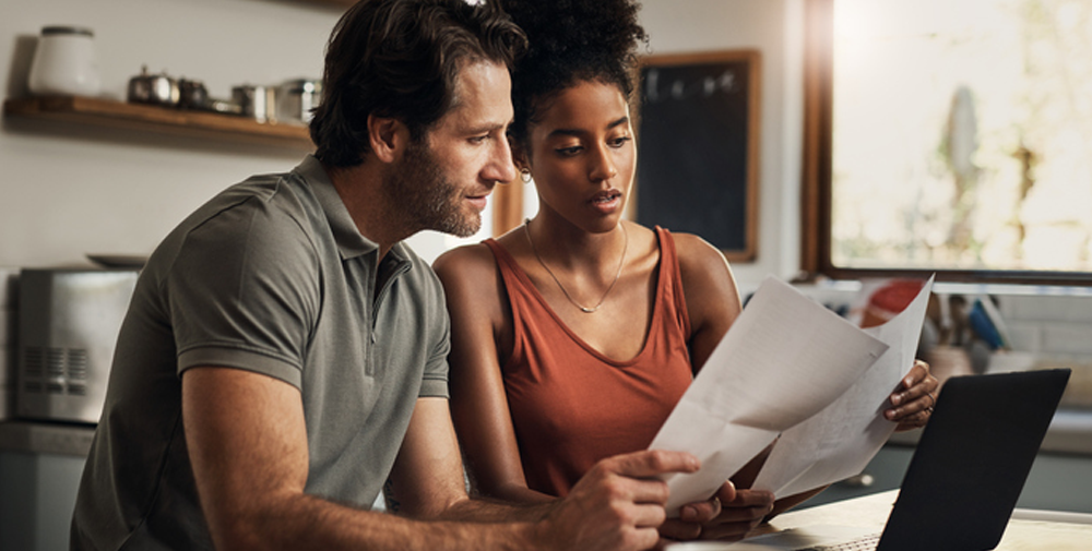 Young couple with a worried look holding two sheets of paper over a laptop in a kitchen.