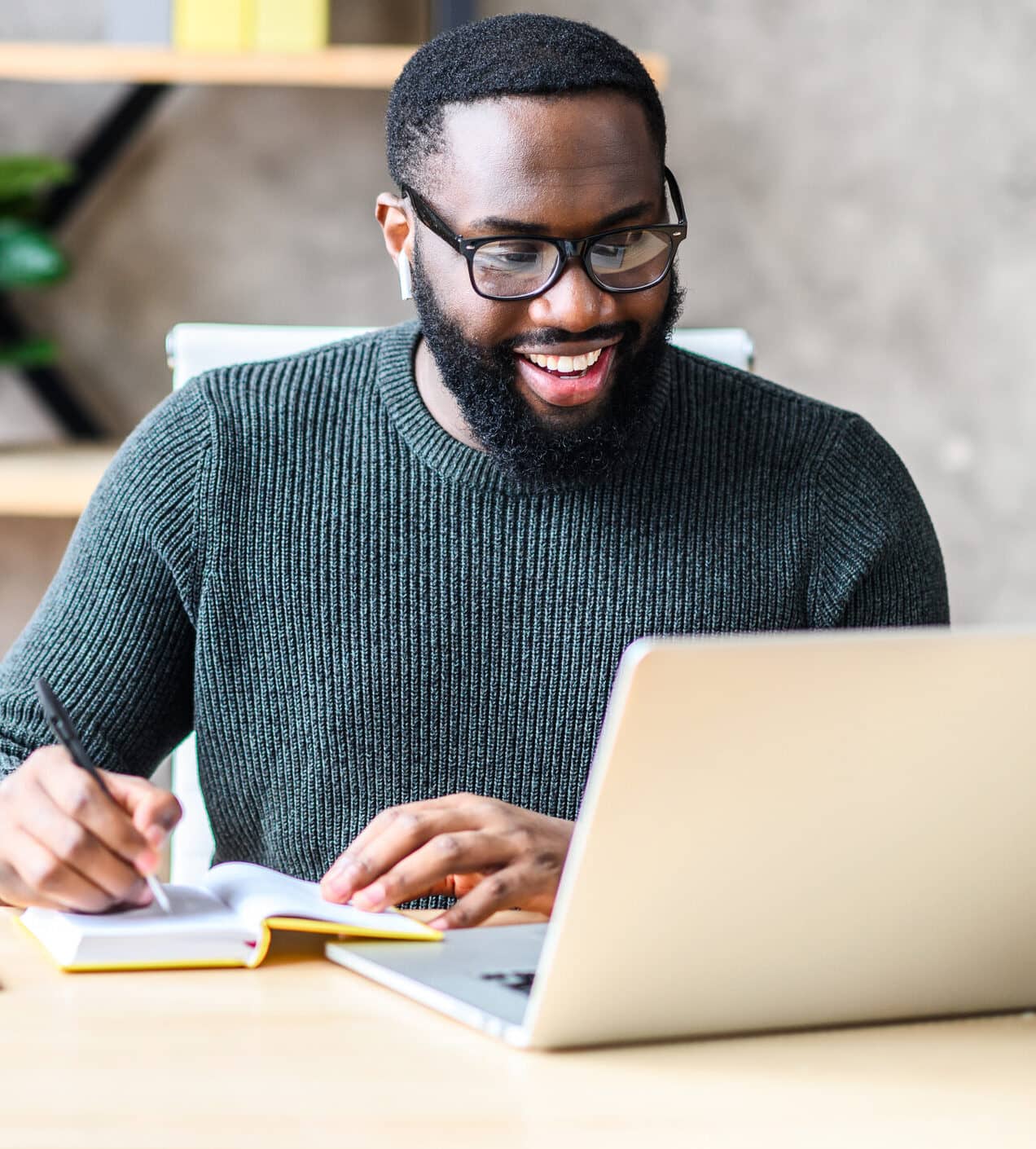 Cheerful African-American male student or worker in glasses is watching online lectures or webinars and writing notes in a notebook