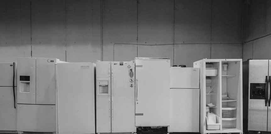 Black and white photo of old refrigerators and freezers lined up against a concrete wall in a warehouse.