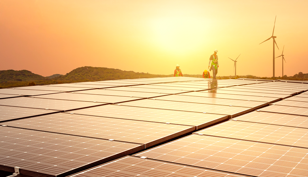 Field of solar panels with windmills and mountains in the background while the sun sets