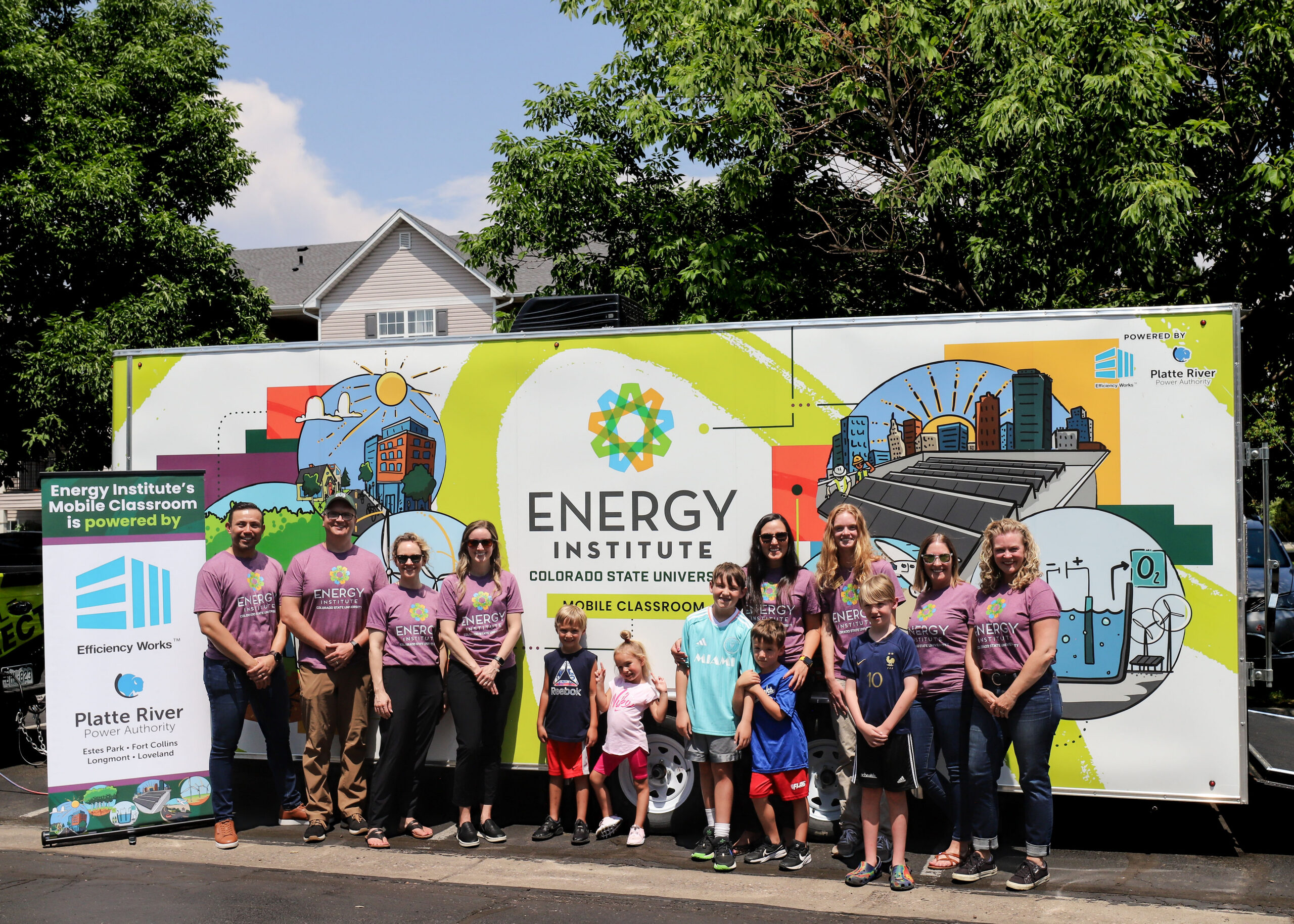 A group of adults and children stand in front of a large trailer decorated with colorful illustrations of buildings, solar panels, and scientific imagery. The trailer displays the logo and name “Energy Institute – Colorado State University Mobile Classroom.” A sign to the left notes that the mobile classroom is powered by Efficiency Works and Platte River. The group is wearing matching purple shirts, and they are standing on a sunny day with trees and houses in the background.
